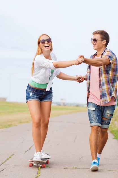 Vakantie, vakantie, liefde en vriendschap concept - glimlachend paar met skateboard rijden en hand in hand buitenshuis