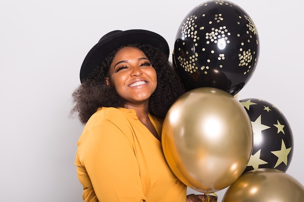 Vakantie, verjaardagsfeestje en leuk concept - Portret van lachende jonge Afro-Amerikaanse jonge vrouw die stijlvol op witte muur met ballonnen kijkt.