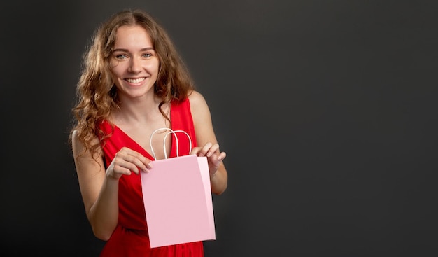 Vakantie verrassing Speciale gelegenheid Gelukkige vrouw in rode jurk met cadeau in merk mockup roze boodschappentas geïsoleerd op donkere kopie ruimte achtergrond Sale korting