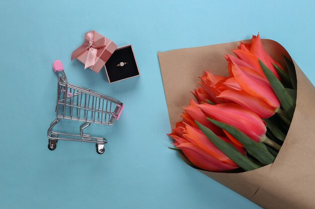 Vakantie winkelen. tulpenboeket, gouden ring met diamant in geschenkdoos, supermarktkarretje op blauw