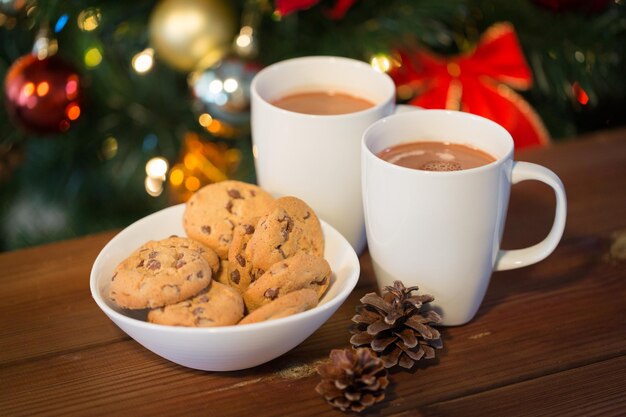 vakantie, winter, eten en drinken concept - close-up van havermoutkoekjes, kopjes met warme chocolademelk of cacaodrank en dennenappels op houten tafel over kerstboomachtergrond