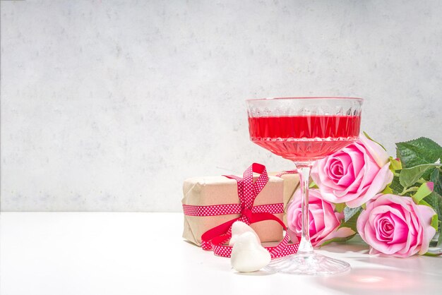 Valentijnsdag, 8 maart, moederdag wenskaart achtergrond. Roze roos bloemboeket, ambachtelijke geschenkdoos met rood lint, rose wijn cocktail op witte tafel achtergrond kopie ruimte