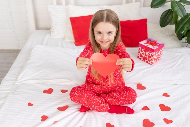 Valentijnsdag concept een schattig kind meisje zit thuis op het bed in rode pyjama's en houdt haar hart in haar handen en glimlachend feliciteert met de vakantie