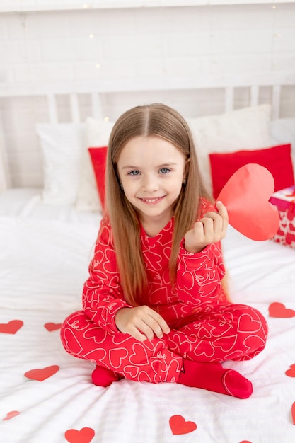 Valentijnsdag concept een schattig kind meisje zit thuis op het bed in rode pyjama's en houdt haar hart in haar handen en glimlachend feliciteert met de vakantie