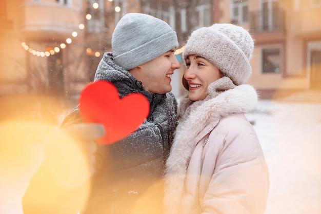 Valentijnsdag concept, liefdevolle paar kus en knuffel in een winter besneeuwde park. Jonge man houdt een hart van rood papier terwijl hij de hele dag met zijn vriendin viert. Een stel dat het samen warm heeft.