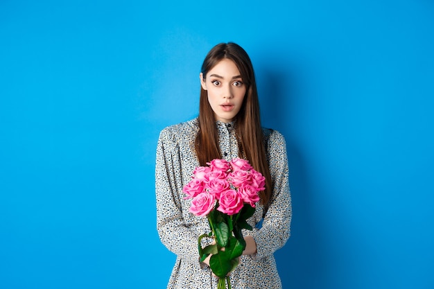 Valentijnsdag concept. Verrast vriendin die een prachtig boeket bloemen ontvangt en met ongeloof naar de camera kijkt, staande op een blauwe achtergrond.