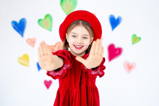 Foto valentijnsdag een klein glimlachend kind een meisje in een rode baret dat gelukkig haar handen zwaait en zich verheugt tegen de achtergrond van kleurrijke harten