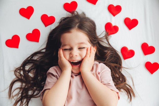 Valentijnsdag een klein meisje dat thuis op een wit bed ligt tussen harten die glimlachen en lachen van geluk feliciteren met de vakantie