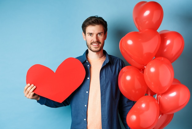 Valentijnsdag en liefde concept. Glimlachende jonge man die minnaar met romantische giften, hartballons en valentijnskaartprentbriefkaar wacht, die zich over blauwe achtergrond bevindt.