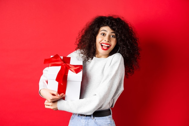 Valentijnsdag. Gelukkig vriendin houden geschenkdoos van geheime aanbidder, romantisch kijken naar minnaar, ontvangen cadeau, staande op rode achtergrond.