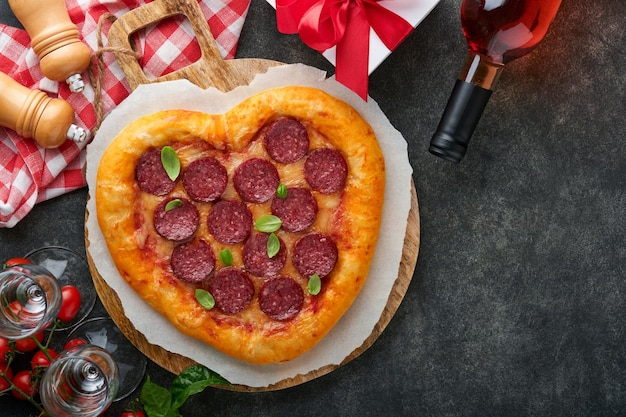 Valentijnsdag hartvormige pizza met mozzarella pepperoni en basilicum wijnfles twee wijnglas geschenkdoos op zwarte achtergrond Idee voor romantisch diner Valentijnsdag Bovenaanzicht Mock up