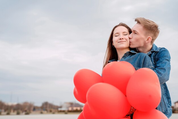 Valentijnsdag. Jong liefdevol paar knuffelen en kussen met rode hartvormige ballonnen buitenshuis