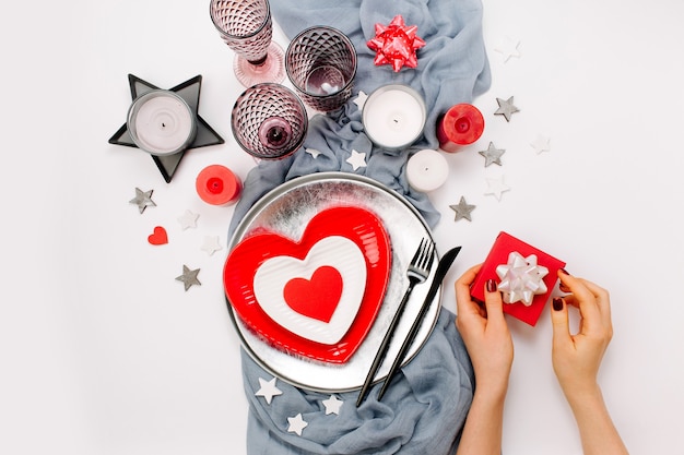 Valentijnsdag tafel instelling. Vrouwelijke handen houden cadeau. Witte en rode gerechten in de vorm van een hart, drankglazen, kaarsen en bestek witte achtergrond. Romantisch concept