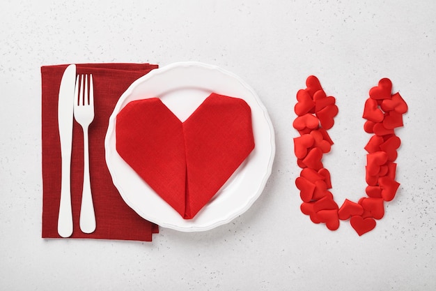 Valentinas day table setting white color with white silverware, red napkin folded as heart on white background for greetings. I love you. Romantic dinner or menu concept. Empty space for text.