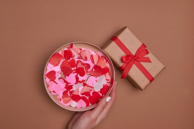 Valentine-dagsamenstelling met exemplaarruimte. Aanwezig of geschenkdoos verpakt in ambachtelijk papier met strik en rode harten. Envelop ansichtkaart met confetti op pastel achtergrond. Close-up, bovenaanzicht, kopieer ruimte