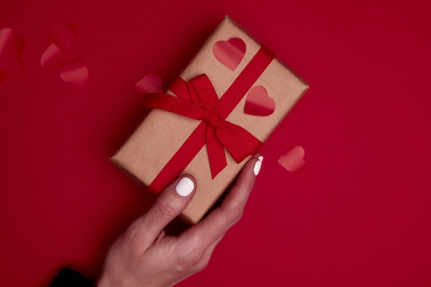 Valentine-dagsamenstelling met exemplaarruimte. Vrouw hand met cadeau of geschenkdoos verpakt in ambachtelijk papier met strik en rode harten confetti. Close-up, bovenaanzicht, kopieer ruimte