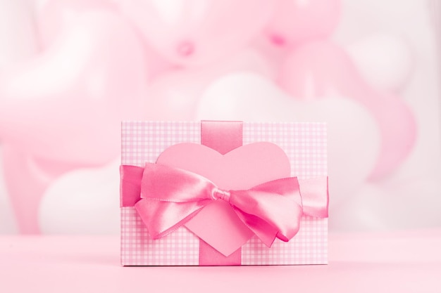 사진 발렌타인 데이 선물은 줄무늬 종이로 싸이고 실크 리본 활과 하트 모양의 인사말 카드로 분홍색 풍선 배경에 텍스트 복사 공간이 있는 상자에 묶여 있습니다.