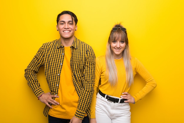 In valentine day young couple over vibrant yellow background posing with arms at hip and smiling