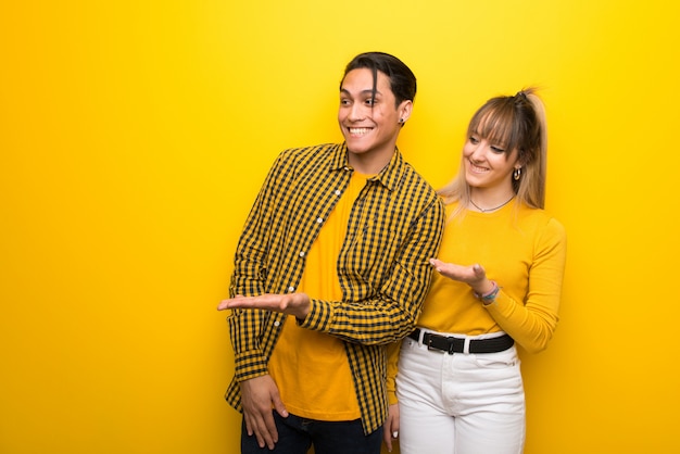 In valentine day Young couple over vibrant yellow background presenting an idea while looking smiling towards