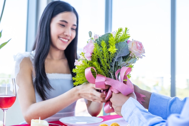 발렌타인 데이 및 아시아 젊은 행복한 커플 개념, 꽃다발 장미를 들고 있는 남자, 그녀의 얼굴에 미소를 짓고 있는 여성에게 손을 얹고 점심 식사 후 놀라움을 기다립니다 레스토랑 배경