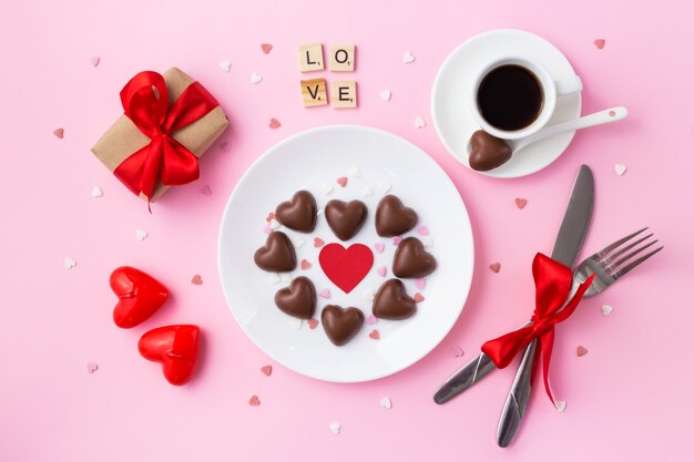 Valentine's day breakfast. Heart made of chocolate candies, gift with a red bow and cu of coffee on pink