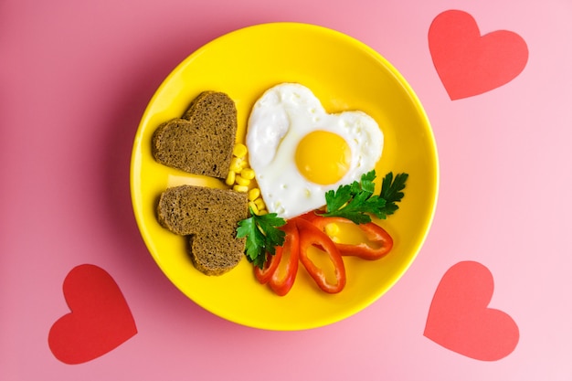День Святого Валентина завтрак. жареное яйцо в форме сердца и хлеб в желтой тарелке на красном
