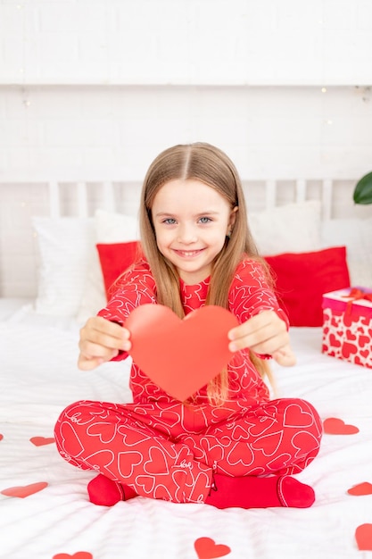 발렌타인 데이 개념 귀여운 소녀가 집에 있는 빨간 잠옷을 입고 침대에 앉아 손에 심장을 잡고 휴가를 축하하며 웃고 있다