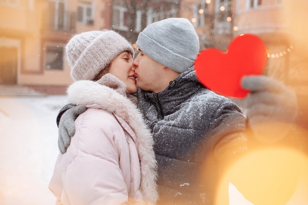 Фото Концепция дня святого валентина, влюбленные поцелуи и объятия в зимнем снежном парке. молодой человек держит красное бумажное сердце, празднуя день всех влюбленных со своей девушкой. пара чувствует тепло вместе.