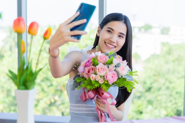 バレンタインのコンセプト、レストランでバラの花束を持ってテーブルフードに座っているアジアの若い女性を笑顔の幸せのSelfie