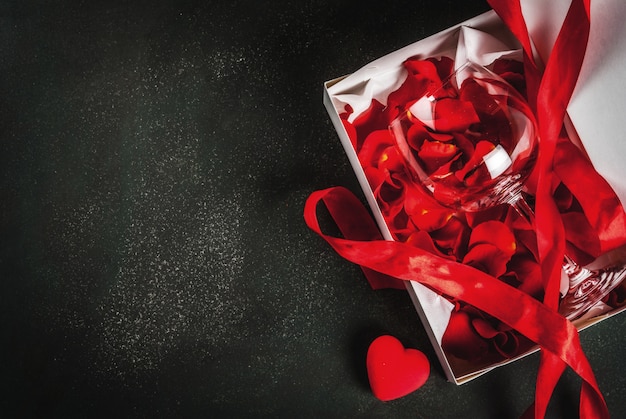 Valentine's day concept, white wrapped gift box with red ribbon, with rose flower petals in wine glass, with red candle, on dark stone background, copy space top view