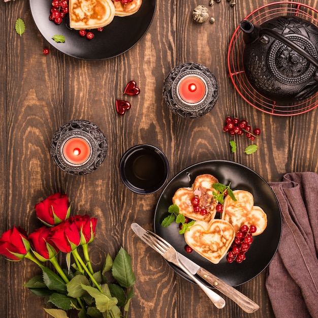 Valentine's day flat lay with delicious pancakes in form of heart, green tea, black teapot, candles and roses.