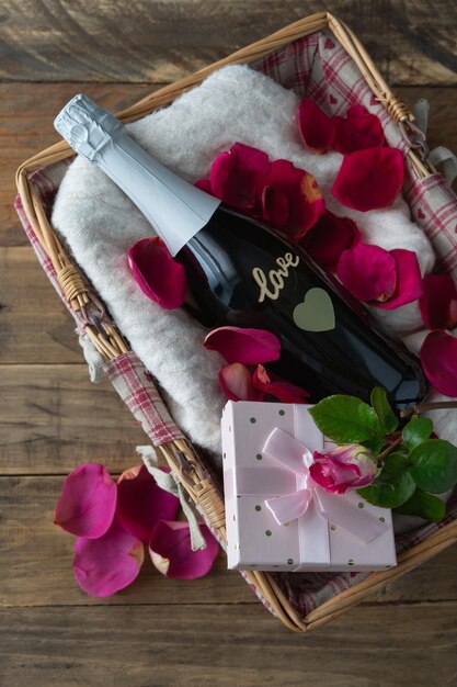 Valentine's Day. Gift basket with champagne bottle with LOVE inscription on the bottle and gift box with rose. Decoration with rose and red petals on wooden background. Copy space.