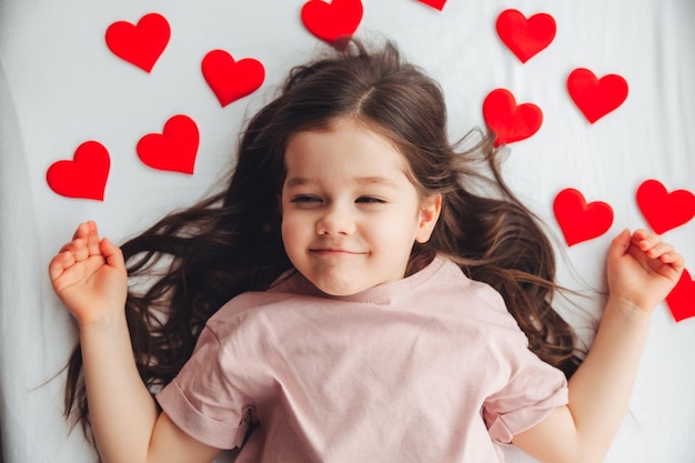 Valentine's Day a little girl lying at home on a white bed among hearts smiling and laughing with happiness congratulating on the holiday