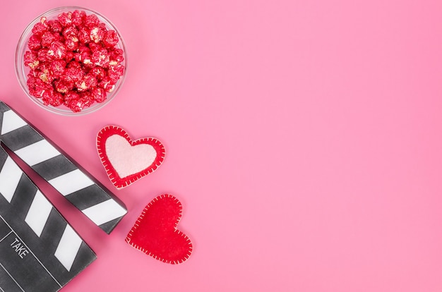 Valentine's Day movie concept. Movie clapperboard with hearts and red caramel popcorn with copy space on pink background.