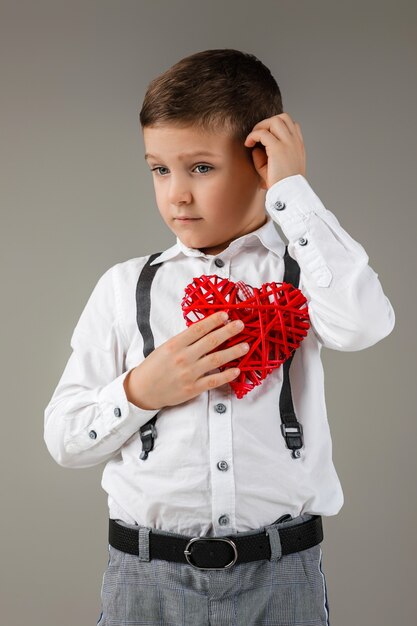 バレンタイン・デー。灰色の背景に分離された赤いハートを持つかわいい子供の男の子の肖像