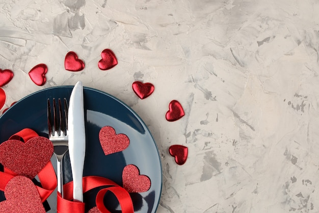 Valentine's Day Table setting Blue plate with a knife and fork and red hearts on a light concrete background.