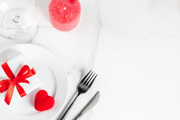 Valentine's day table setting with plate, gift box and red heart, on white marble scene top view 
