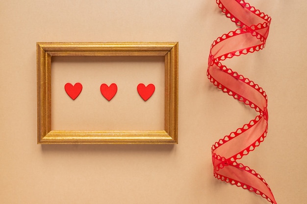 Valentine's day or Wedding romantic concept. Twisted decorative ribbon and golden photo frame with red hearts on beige background.