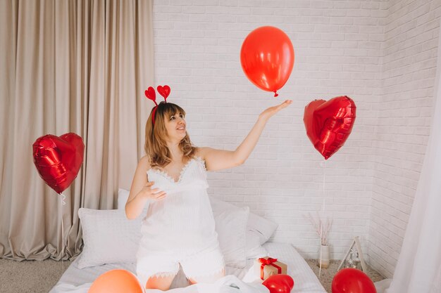 Valentine's Day, Women's Day. Young caucasian girl sitting in bed and celebrating valentine's day holding a heart-shaped balloon in her hands