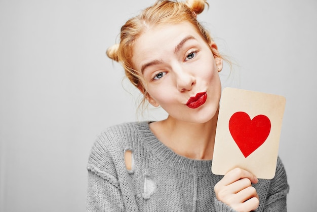 Valentine's day. Young girl in sweater holding a card with a heart. On a gray background