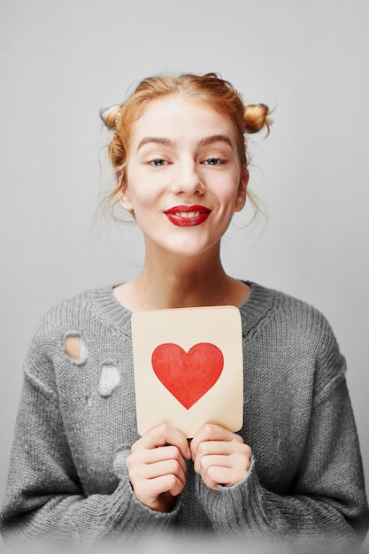 Valentine's day. Young girl in sweater holding a card with a heart. On a gray background