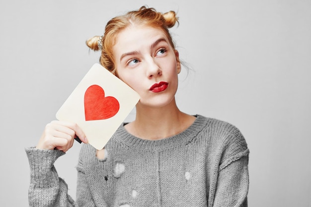 Valentine's day. Young girl in sweater holding a card with a heart. On a gray background