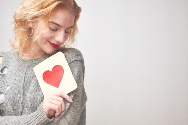 Valentine's day. Young girl in sweater holding a card with a heart. On a gray background