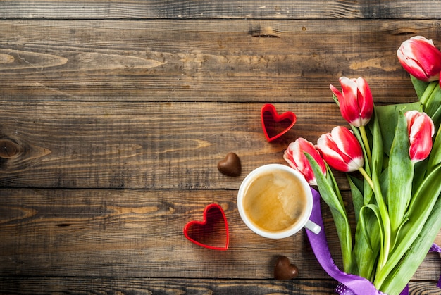 おめでとう、グリーティングカードのバレンタインデーの背景。木製の背景平面図コピースペースにチョコレートハートキャンディとコーヒーマグカップと赤の心で新鮮な春のチューリップの花