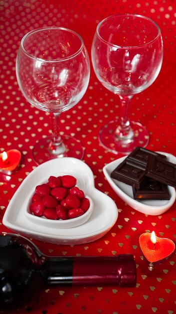 San valentino. bottiglia di vino, bicchieri, rose rosse, candele - sfondo rosso. concetto di cena d'amore - foto verticale