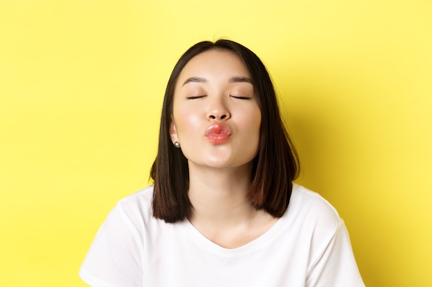 Valentines day concept. Close up of cute asian girl pucker lips and close eyes for kiss, standing romantic against yellow.