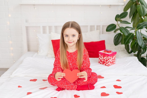 발렌타인 데이 개념 귀여운 소녀가 집에 있는 빨간 잠옷을 입고 침대에 앉아 손에 심장을 잡고 휴가를 축하하며 웃고 있습니다