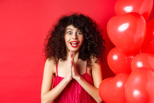 Valentines day. Excited caucasian woman in dress jumping from amazement, looking at something taunting, want to get product, standing near hearts balloons, white background.