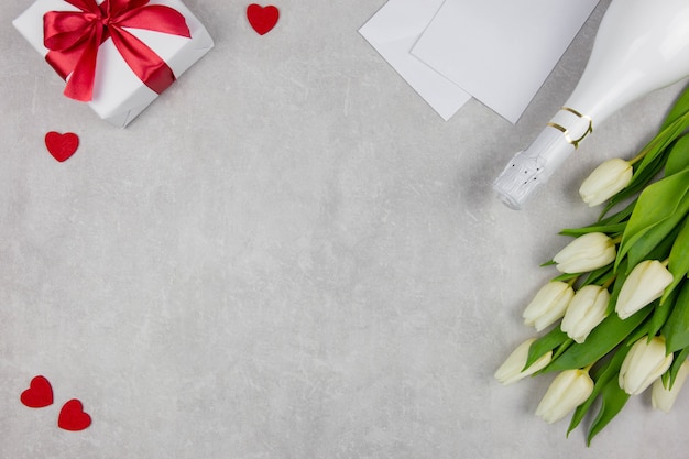 Valentines day flat lay with champagne, gift box, red hearts and white tulips flowers bouquet on light concrete.