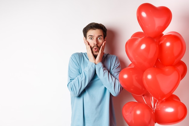 Valentines day. Image of young man standing near hearts balloons with shocked face, staring startled at camera, white background.